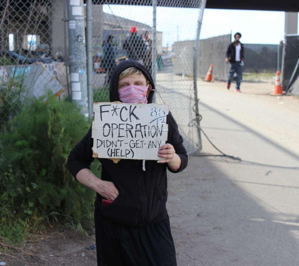 Jessica Bailey, who is unhoused, poses at Wood St, where she currently lives, with a sign she brought to the protest outside of Mayor Libby Schaaf's home on Monday. Her sign criticizes Operation Dignity, the non-profit who has contracted with the city of Oakland to run the Tuff Shed site that Baily lived in for over three months. Photo by Zack Haber.