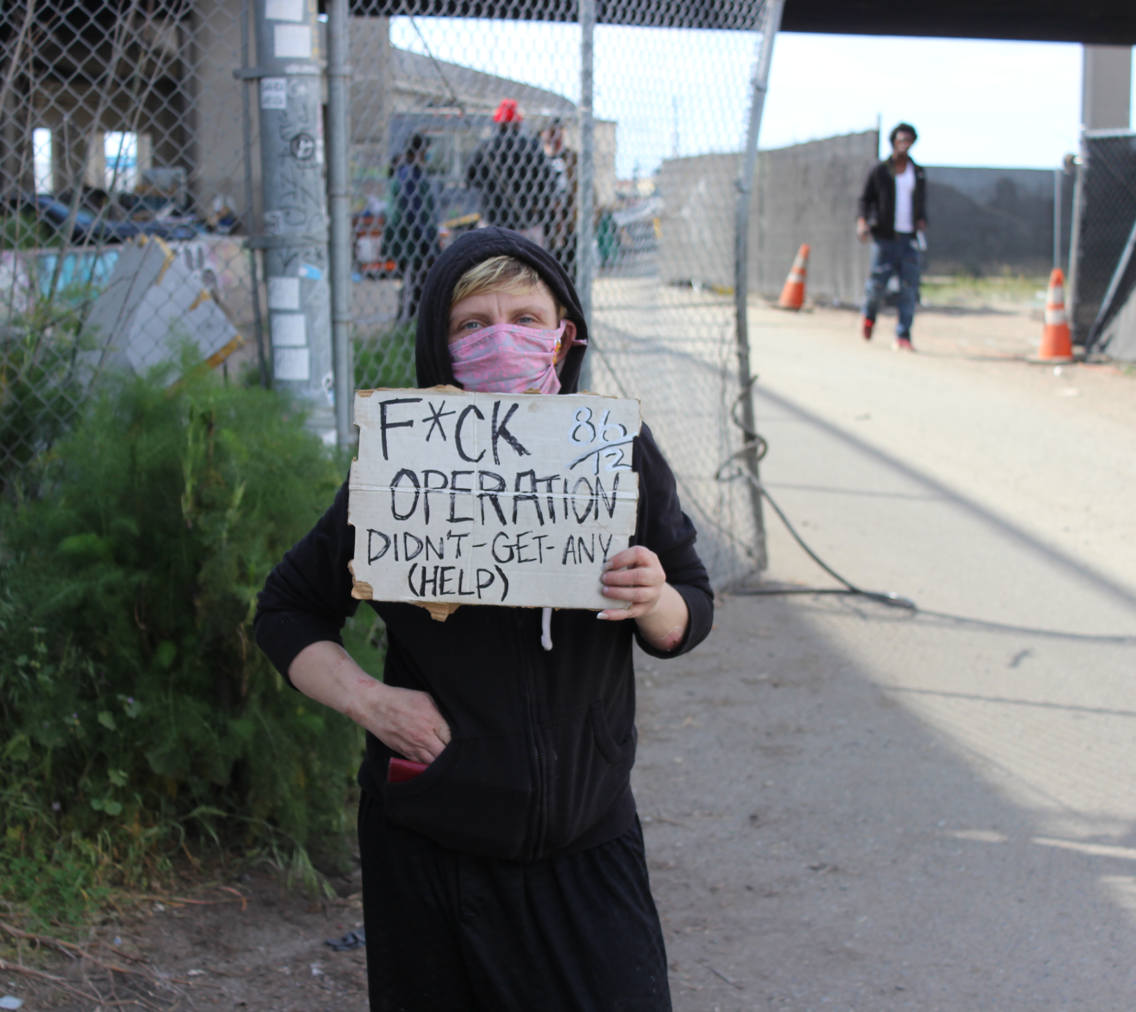 Jessica Bailey, who is unhoused, poses at Wood St, where she currently lives, with a sign she brought to the protest outside of Mayor Libby Schaaf's home on Monday. Her sign criticizes Operation Dignity, the non-profit who has contracted with the city of Oakland to run the Tuff Shed site that Baily lived in for over three months. Photo by Zack Haber.